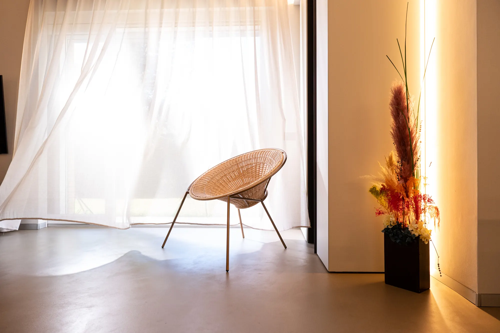 Poltroncina intrecciata davanti a una tenda, con fiori secchi in un vaso nero