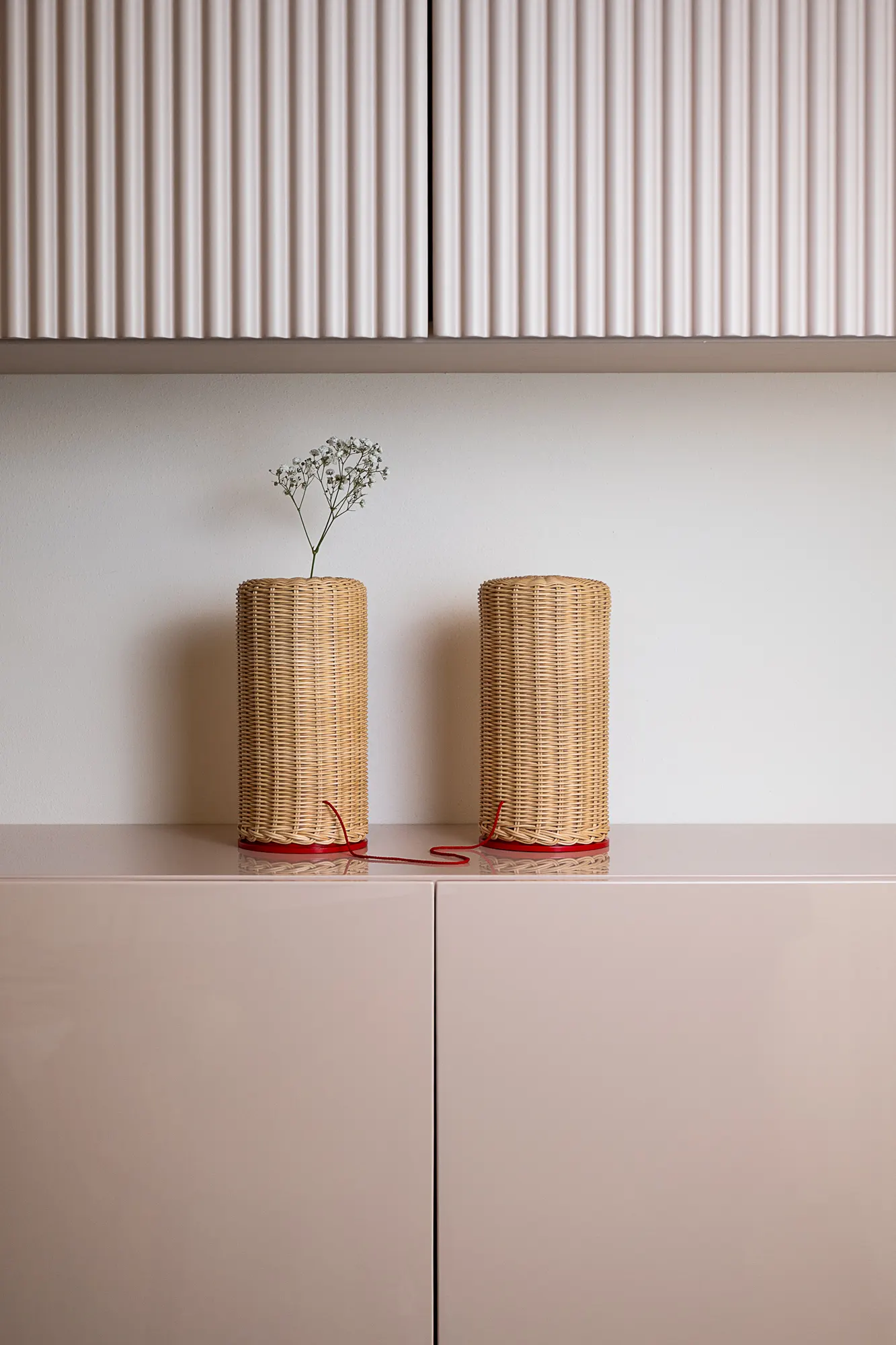 Due lampade cilindriche in midollino su una credenza rosa, ante a doghe verticali