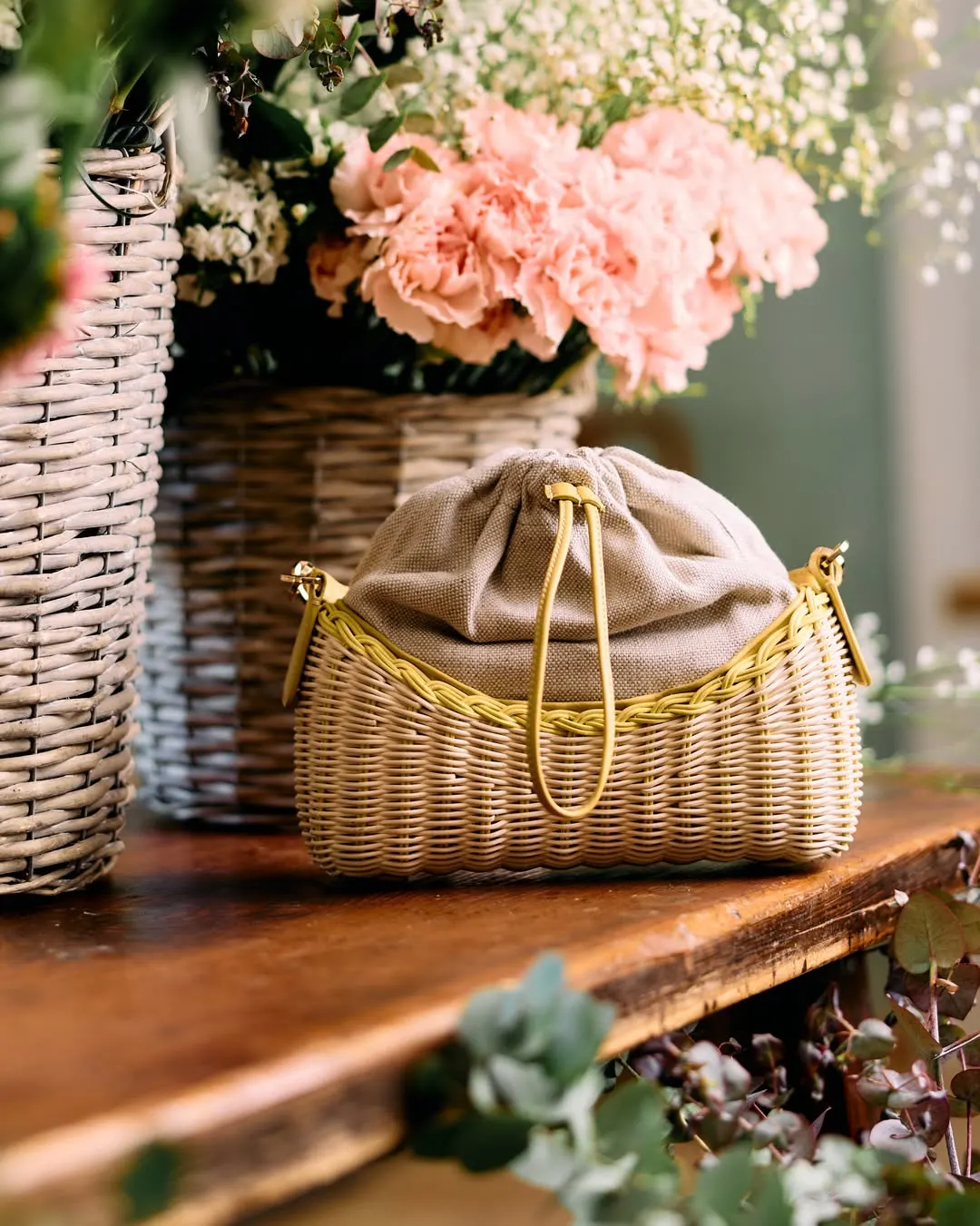 Borsa a sacchetto in paglia con catena dorata accanto a un cesto di fiori rosa